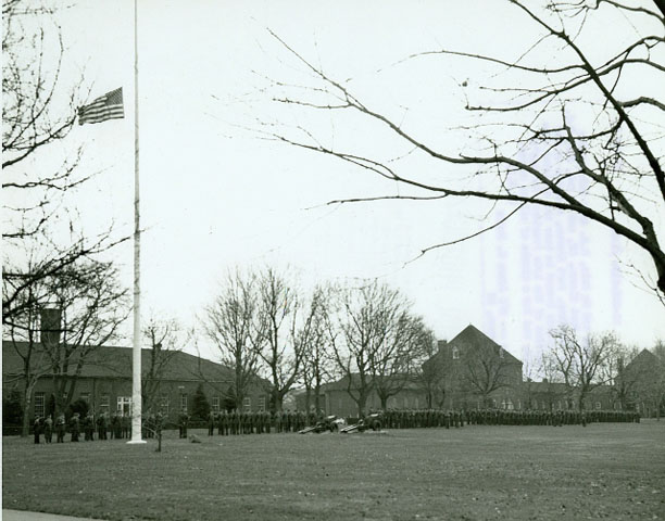 <p>Personnel in formation at the post flagpole marking the death of President John F. Kennedy on November 22, 1963. Looking southeast toward the Mess Hall (Building 67, left) and Barracks (Building 68, right).</p>