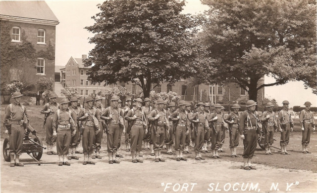 <p>Taken in the spring of 1942, this photo postcard may show the combat platoon organized early in the Second World War to protect Fort Slocum from saboteurs and commandos.&#160; The men stand south of Buildings 61 (left) and 59-60 (right).&#160; With equipment spanning 30-plus years of Army designs—from 1903 Springfield rifles through 1917 Brodie helmets and water-cooled machine guns (on carts) to 1930s uniforms—the platoon embodies the American soldier in transition from doughboy to GI.</p>