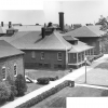 <p>Pair of photos joined together to show the western side of the Barracks Area, with the Guardhouse (Building 56) at center and portions of one of the barracks (Building 55) and the Mess Hall (Building 67) (left-right) to either side. View southeast, mid 1950s.</p>